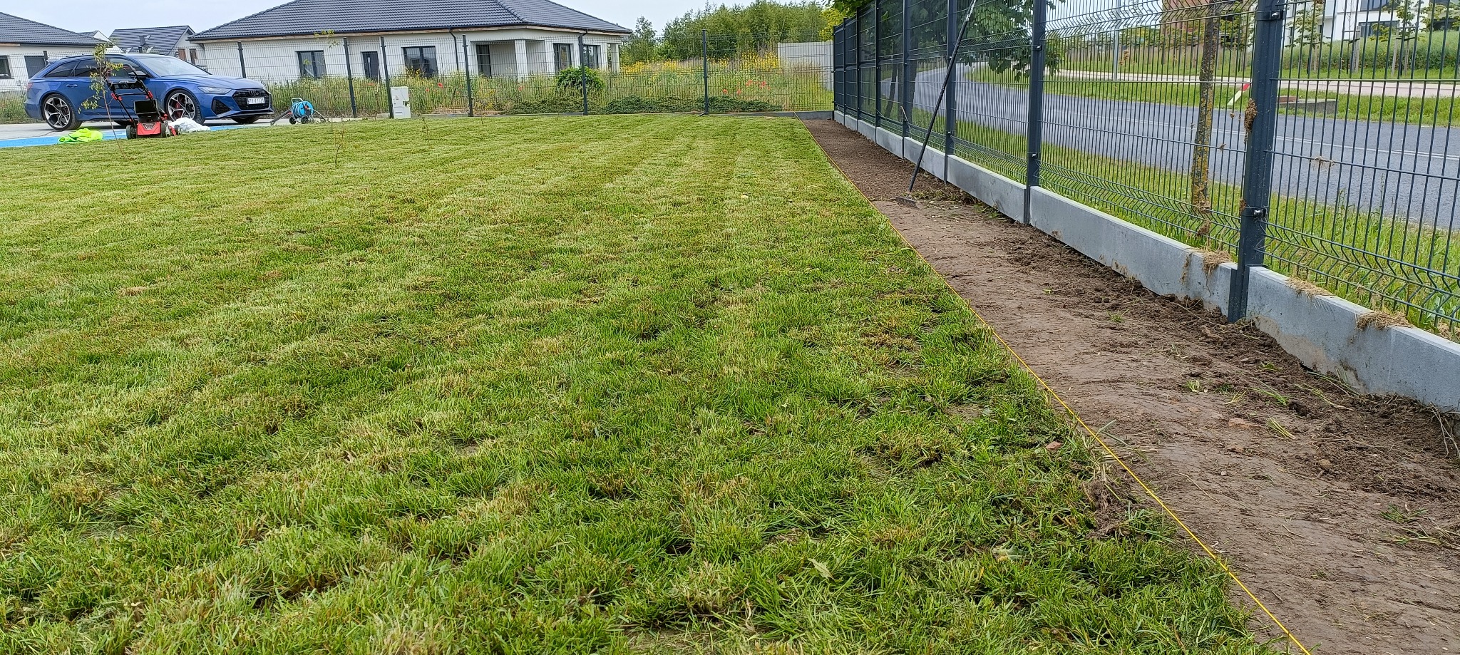 Świeżo założony trawnik z rolki, widoczna granica trawy i przygotowane obrzeże wzdłuż ogrodzenia. W tle dom i samochód.
