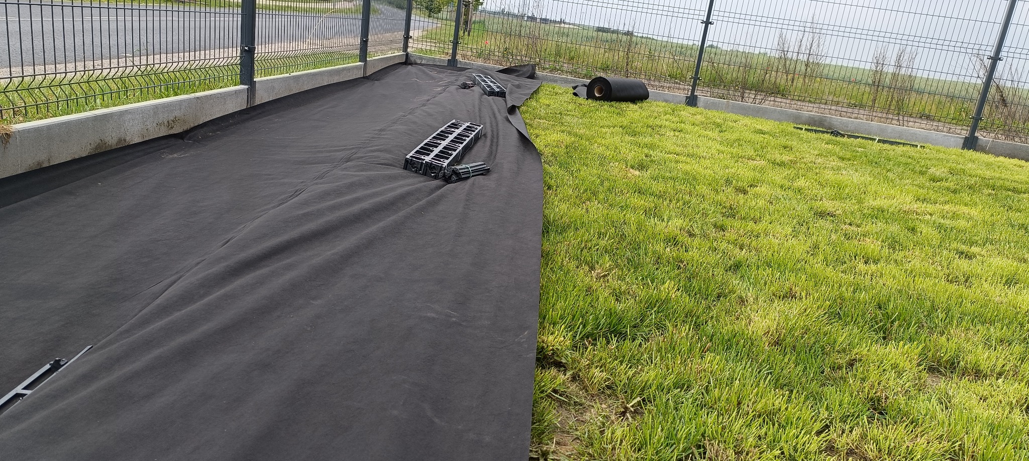 Rozłożona czarna agrowłóknina z kotwami na trawniku, przygotowanie pod trawnik z rolki. W tle rolka materiału i metalowe ogrodzenie.