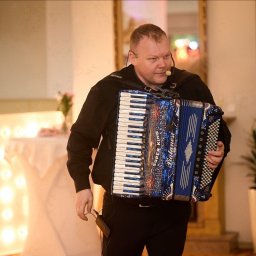 MARTEX MARCIN GAJEWSKI - Akordeonista z mikrofonem nagłownym, w trakcie występu na tle dekoracji świetlnych i roślin doniczkowych. Widoczny ozdobny akordeon.
