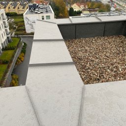 Bagley - Płaski dach pokryty szarą blachą z widocznymi kroplami deszczu i żwirem na obrzeżach, widok z góry na budynki mieszkalne w tle.