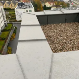 Płaski dach pokryty szarą blachą z widocznymi kroplami deszczu i żwirem na obrzeżach, widok z góry na budynki mieszkalne w tle.