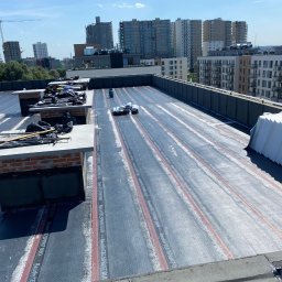 Bagley - Widok z góry na płaski dach w trakcie prac dekarskich, z widocznymi rolkami papy termozgrzewalnej BauderTEC SPRINT DUO, kominami z cegły i panoramą miasta w tle.