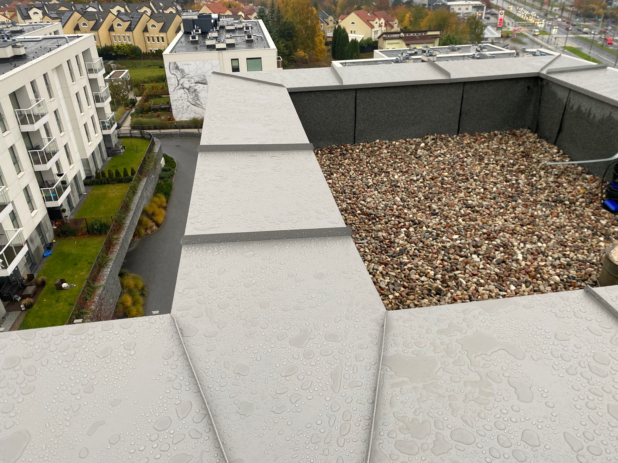Płaski dach pokryty szarą blachą z widocznymi kroplami deszczu i żwirem na obrzeżach, widok z góry na budynki mieszkalne w tle.