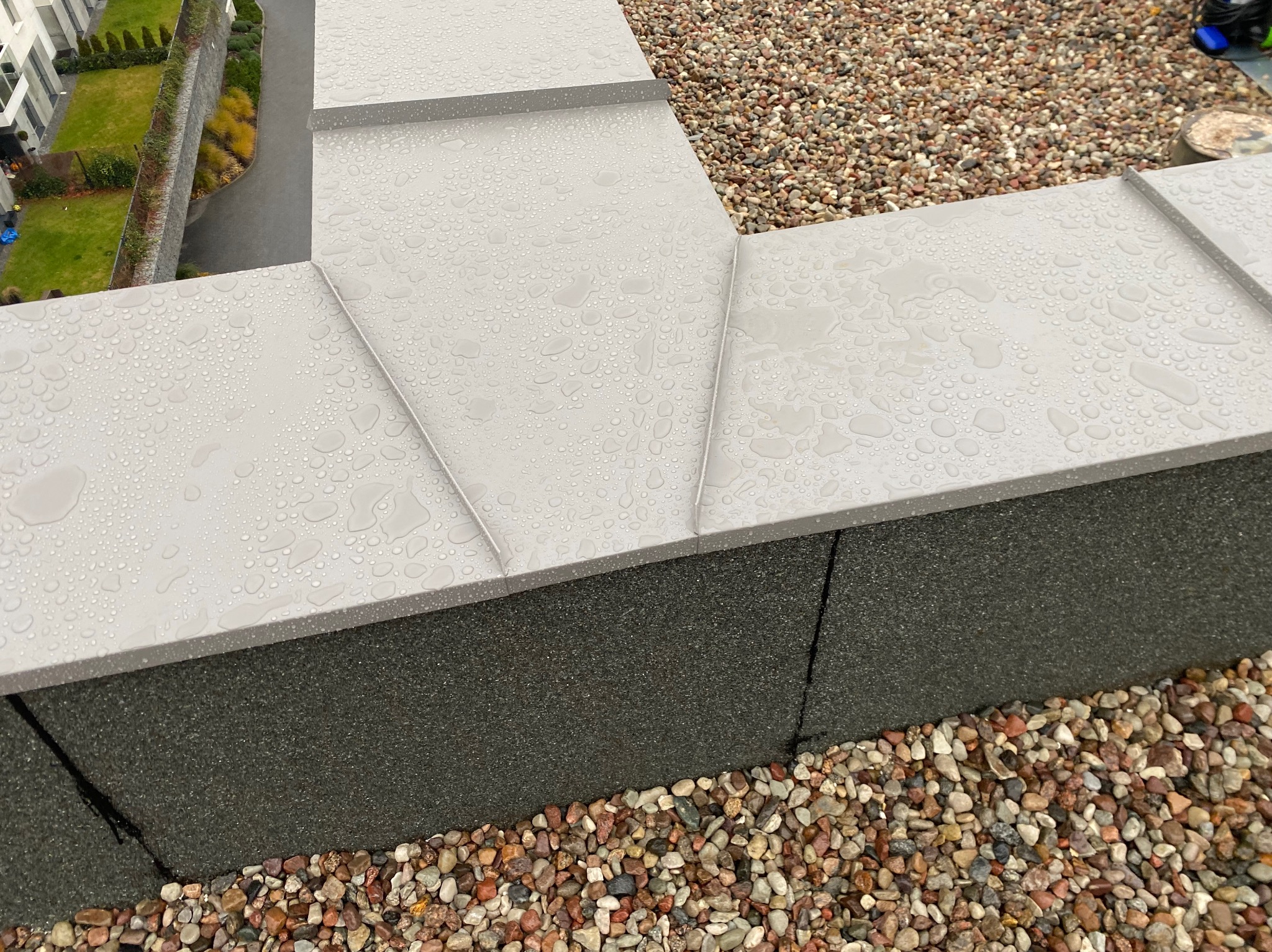 Szary dach pokryty kroplami deszczu, widoczne obróbki blacharskie i kamyczki na dachu, w tle budynek mieszkalny z zielenią.