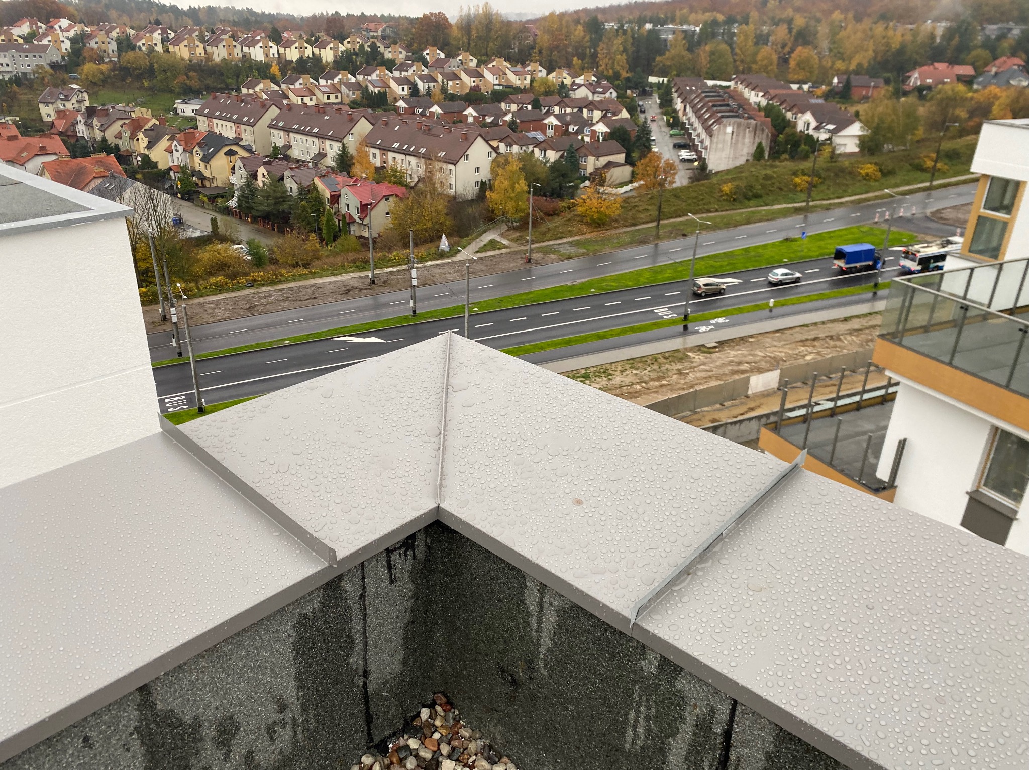 Szare, metalowe obróbki blacharskie na dachu budynku wielorodzinnego, widoczne krople deszczu i fragmenty elewacji. W tle zabudowa miejska i jesienne drzewa.