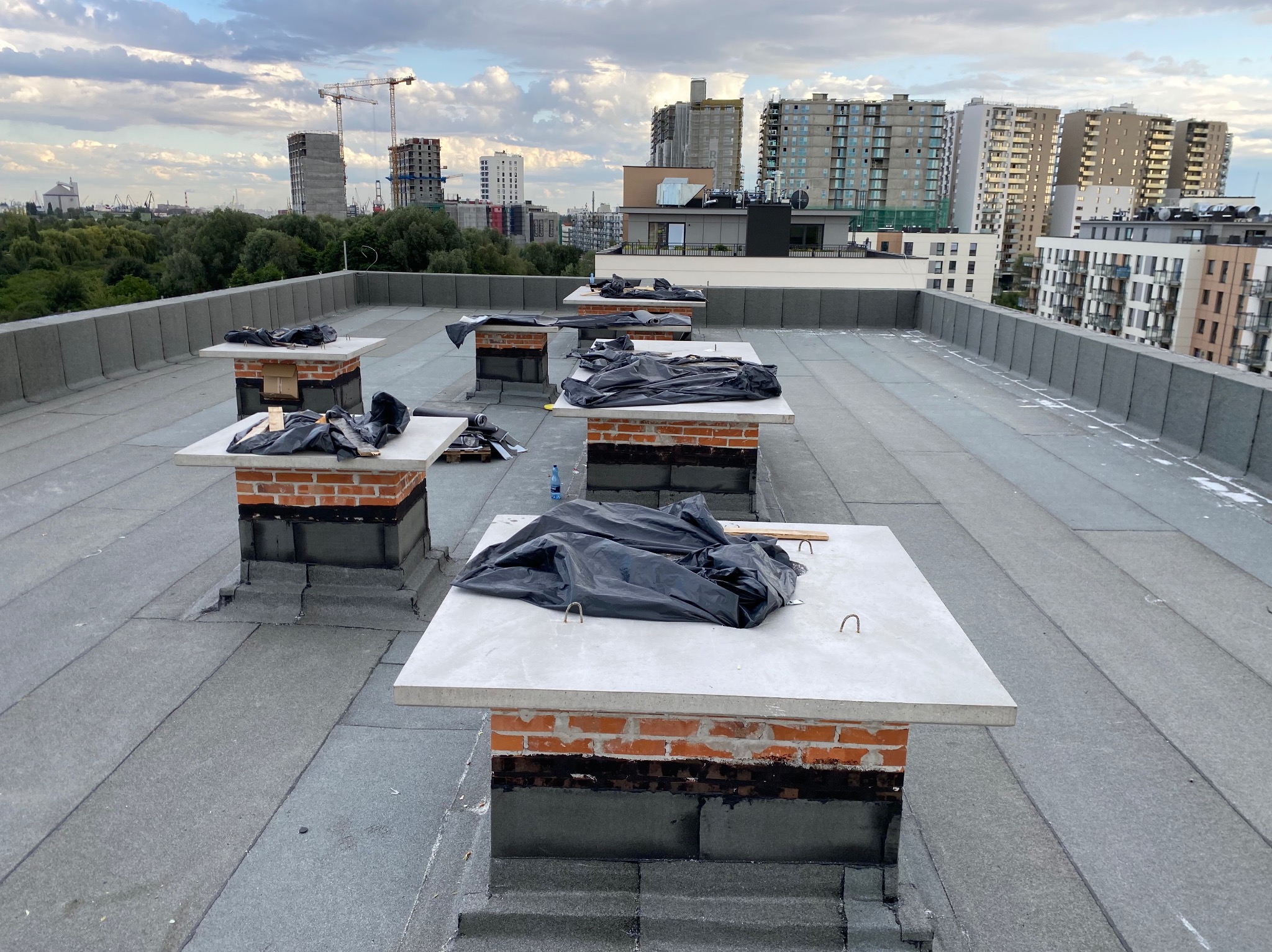 Szary dach pokryty papą termozgrzewalną z kilkoma kominami obłożonymi cegłą i przykrytymi betonowymi płytami, zabezpieczonymi czarną folią, w tle widok na miasto i budowę.