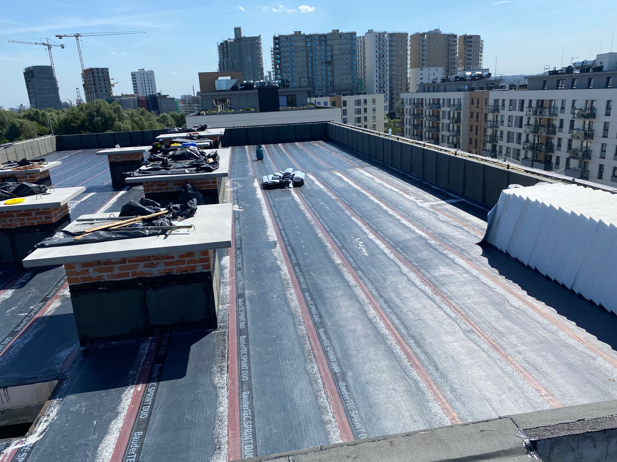 Widok z góry na płaski dach w trakcie prac dekarskich, z widocznymi rolkami papy termozgrzewalnej BauderTEC SPRINT DUO, kominami z cegły i panoramą miasta w tle.