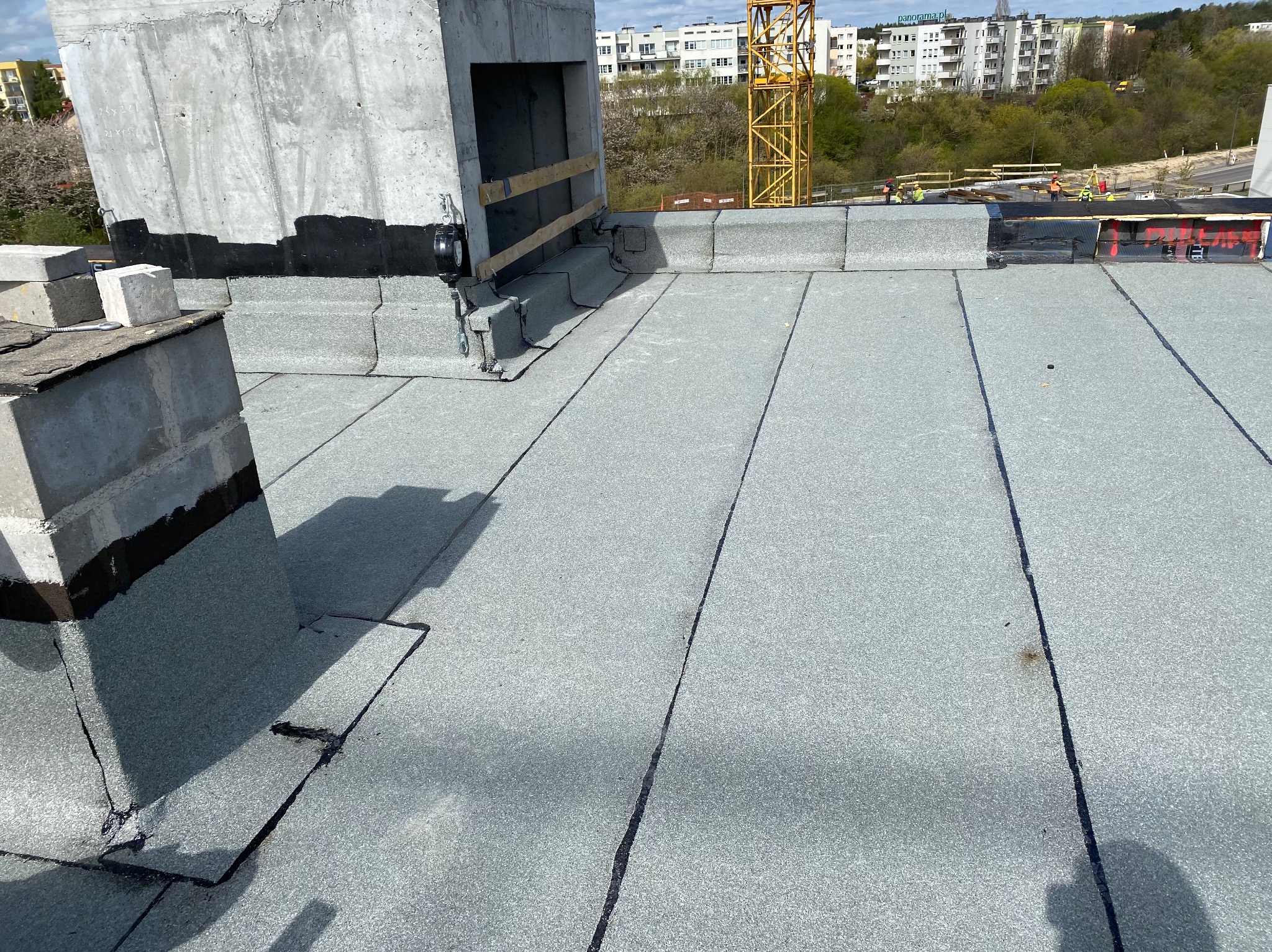 Szary, płaski dach pokryty papą termozgrzewalną z widocznymi łączeniami, komin z betonu zbrojonego zabezpieczony czarną izolacją, w tle budowa i żuraw.
