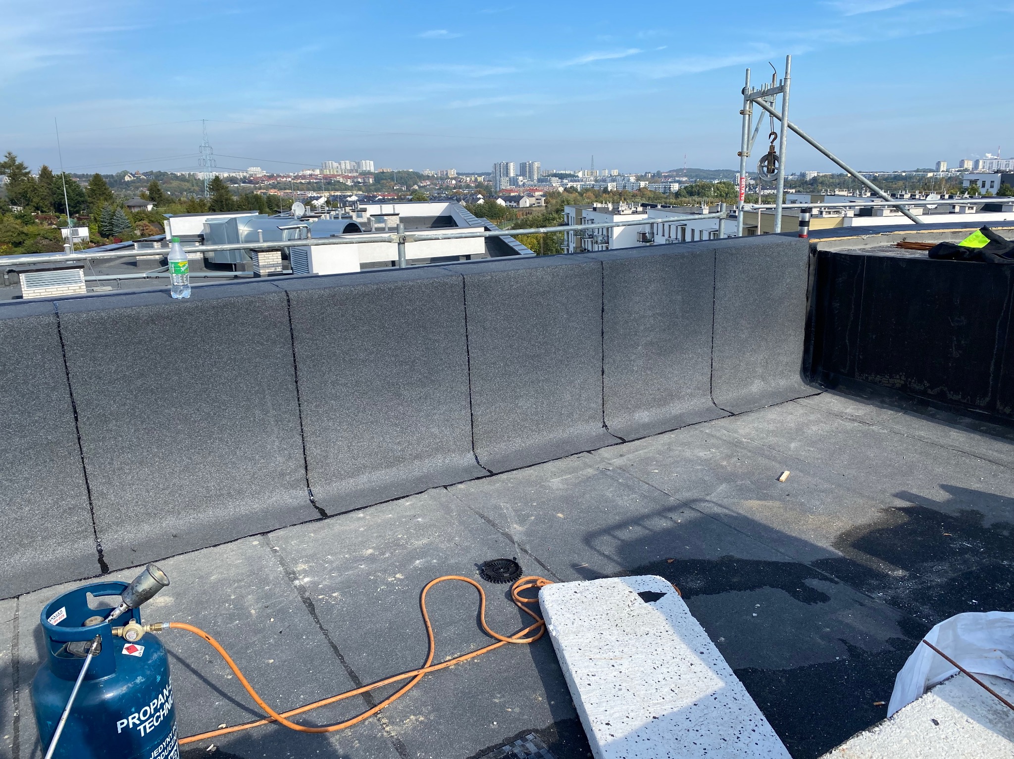 Niski dach pokryty papą termozgrzewalną z widocznym sprzętem dekarskim, butlą gazową i palnikiem, w tle panorama miasta.