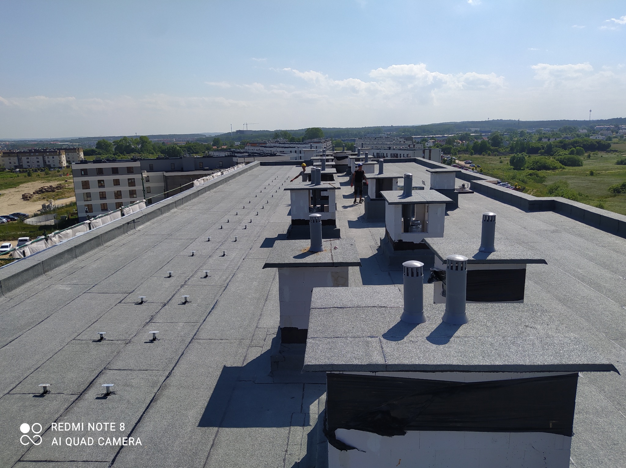 Rozległy, płaski dach budynku wielorodzinnego z szarą papą, licznymi kominami wentylacyjnymi i dwoma pracownikami w oddali, na tle zabudowy miejskiej i zieleni.