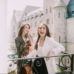 Dwie uśmiechnięte kobiety na balkonie z widokiem na budynek w stylu zamkowym, jedna trzyma wiolonczelę, druga opiera się o balustradę, obie ubrane w eleganckie marynarki.