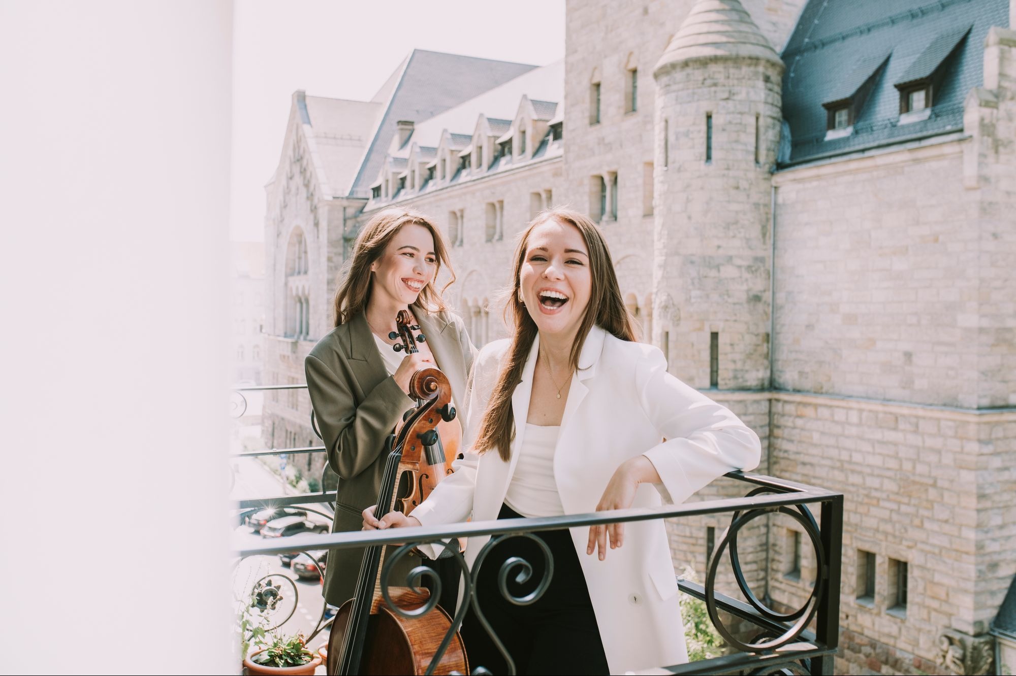 Dwie uśmiechnięte kobiety na balkonie z widokiem na budynek w stylu zamkowym, jedna trzyma wiolonczelę, druga opiera się o balustradę, obie ubrane w eleganckie marynarki.