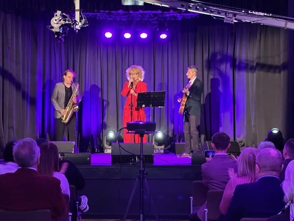 Trójka muzyków na scenie podczas występu: wokalistka w czerwonym stroju śpiewa do mikrofonu, saksofonista w szarej marynarce gra na saksofonie, a gitarzysta w ciemnej marynarce gra na gitarze...