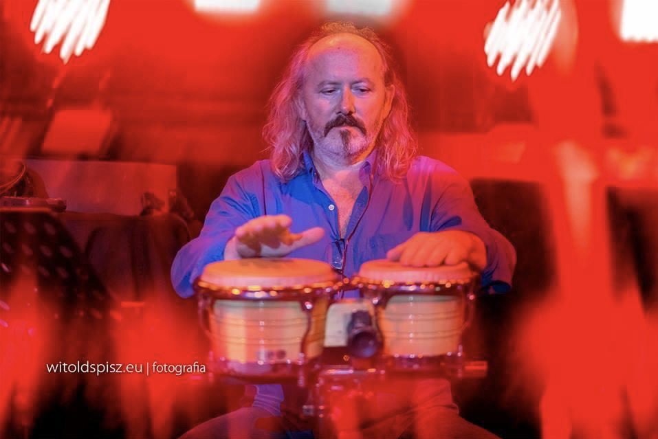 Muzyk z długimi włosami i brodą gra na bębnach bongo w czerwonym oświetleniu scenicznym, skupiony i zaangażowany w występ.