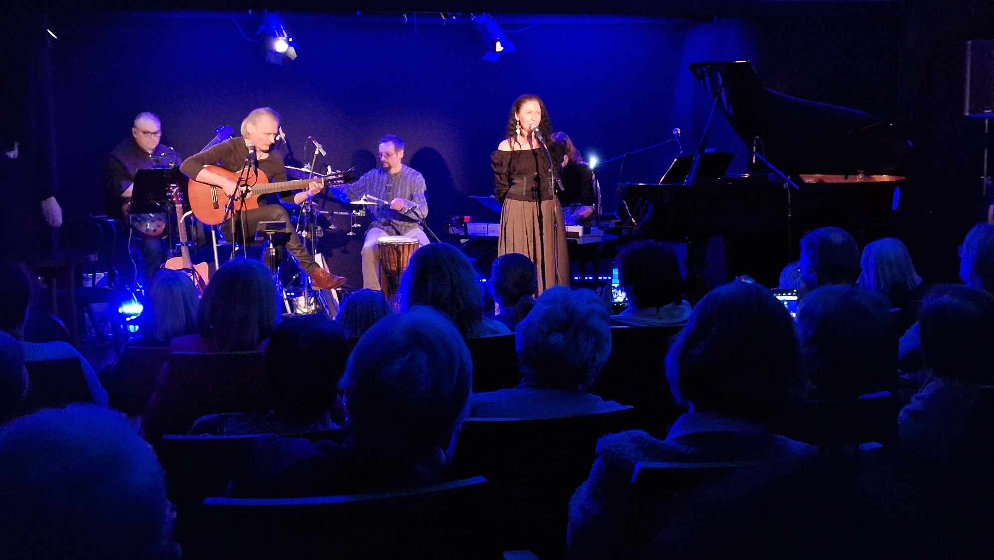 Wokalistka w długiej spódnicy i bluzce z odkrytymi ramionami śpiewa na scenie, obok gitarzysta i perkusista grający na bębnie djembe, w tle czarny fortepian, widownia w ciemnościach.