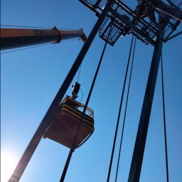 Michał Wysiecki - Usługi Elektryczne Smart Home - Praca na wysokości: Elektryk w koszu dźwigu, montaż instalacji odgromowej na konstrukcji stalowej. Widok z dołu, błękitne niebo w tle.