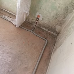 Michał Wysiecki - Usługi Elektryczne Smart Home - Szare rury instalacji elektrycznej ułożone wzdłuż ściany w surowym wnętrzu. Widoczne gniazdko elektryczne w ścianie i fragment zasłony w oknie. Noga w bucie na niebieskiej folii.