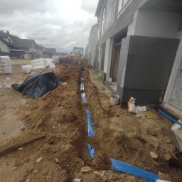 Michał Wysiecki - Usługi Elektryczne Smart Home - Wykop z niebieskimi rurami w ziemi, prawdopodobnie pod instalacje elektryczne, na tle budowy domu i koparki. Widok z góry, pochmurne niebo.
