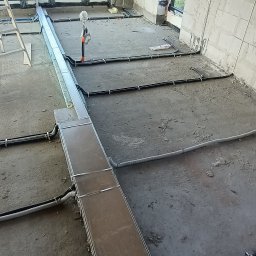 Michał Wysiecki - Usługi Elektryczne Smart Home - Wnętrze w trakcie prac: widoczne kable elektryczne i metalowe korytka kablowe ułożone na surowym betonie, duże okna z widokiem na zewnątrz, instalacja w trakcie realizacji.