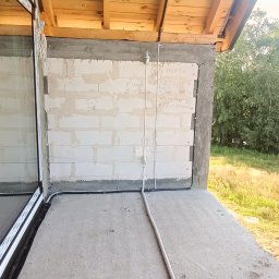 Michał Wysiecki - Usługi Elektryczne Smart Home - Surowy stan budynku z widoczną instalacją elektryczną na ścianie z bloczków betonowych i pod drewnianym stropem. Widoczne kable i rury instalacyjne.