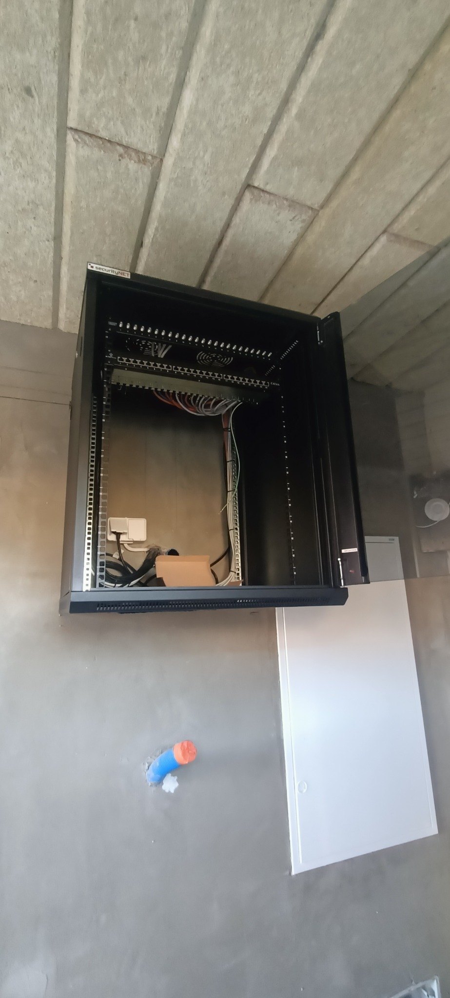 Czarna szafa rack zamontowana na szarej ścianie, widoczne kable i elementy instalacji. Sufit betonowy, w tle biała skrzynka. Ujęcie z dołu.