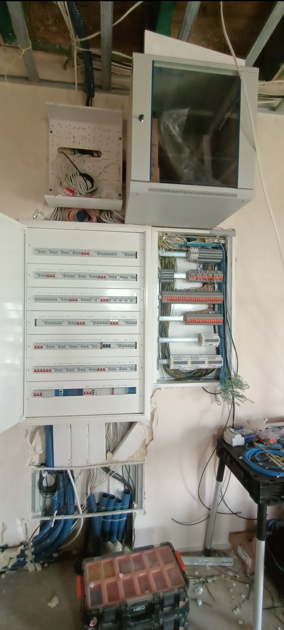 Rozdzielnia elektryczna z otwartymi drzwiczkami, widoczne kable i zabezpieczenia. Dodatkowo skrzynka narzędziowa NEO na podłodze.