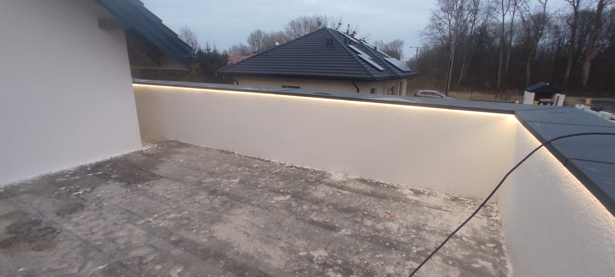 Biała ściana z subtelnym oświetleniem LED na zewnątrz budynku. Widoczne elementy dachu i otoczenia. Ujęcie z dołu, podkreślające linię światła.