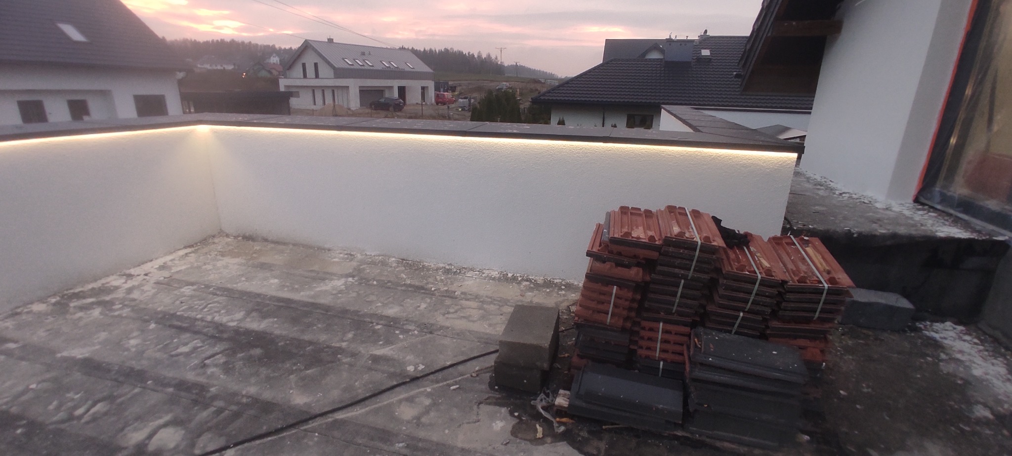 Biała ściana z oświetleniem LED na zewnątrz budynku, stosy dachówek w tle, widok na domy w oddali, wieczorne niebo.