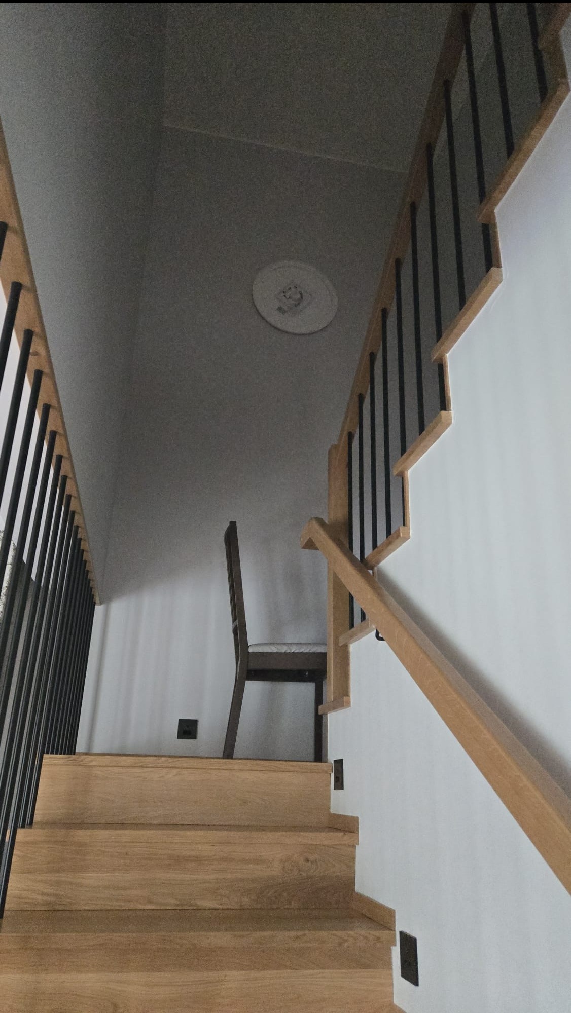 Drewniane schody z czarnymi balustradami, widok z dołu. Na podeście stoi krzesło. Minimalistyczny design, jasne wnętrze.