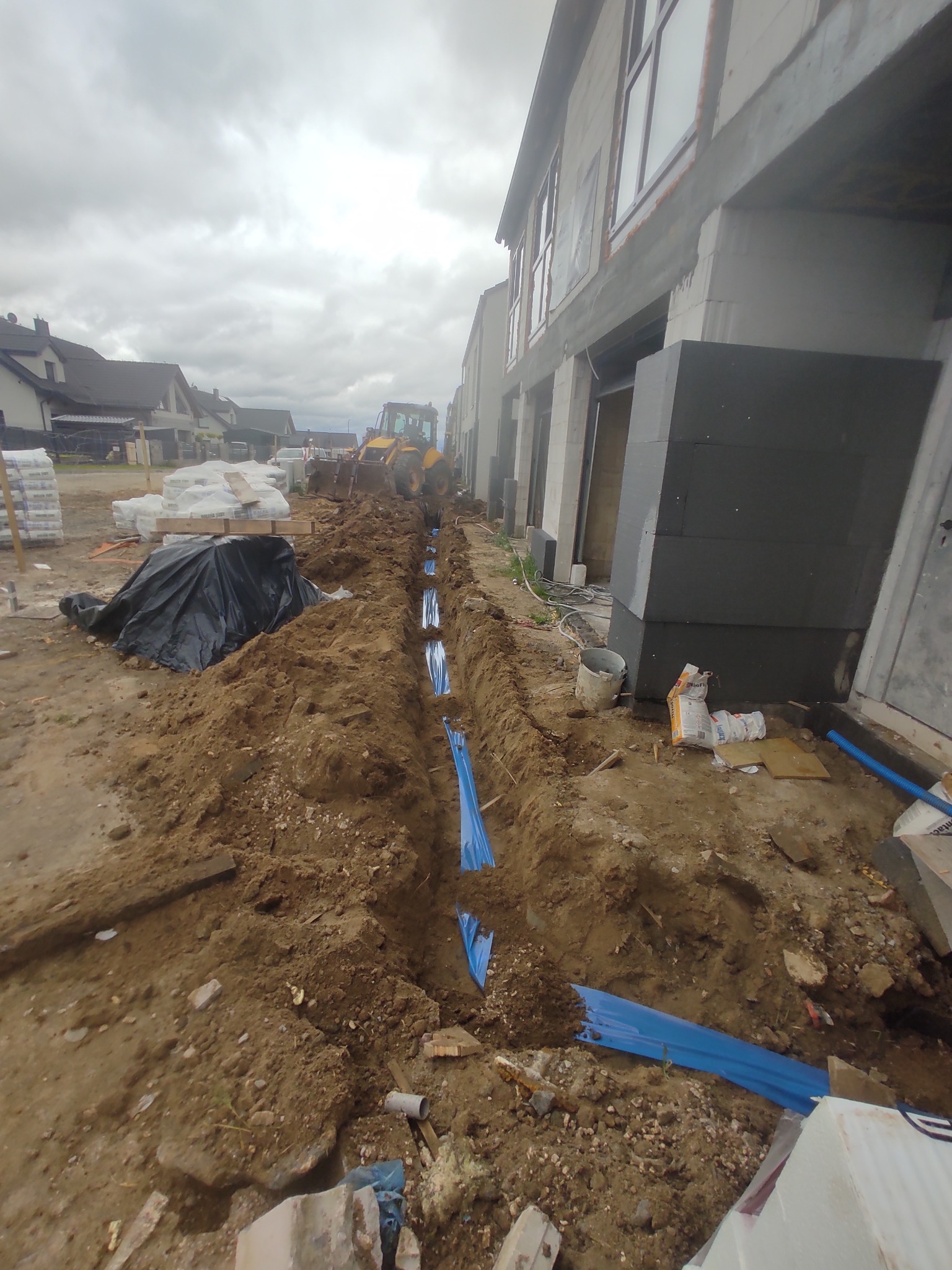 Wykop z niebieskimi rurami w ziemi, prawdopodobnie pod instalacje elektryczne, na tle budowy domu i koparki. Widok z góry, pochmurne niebo.