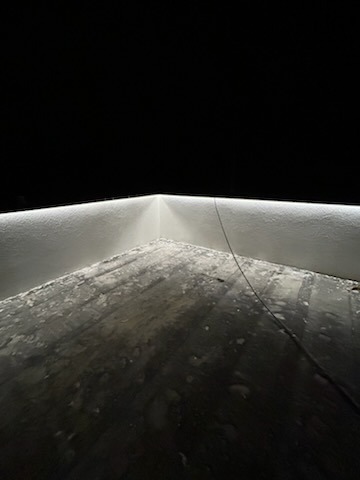 Kąt pomieszczenia z zamontowanym oświetleniem LED, emitującym jasne, białe światło. Widoczny kabel zasilający. Podłoga w stanie surowym, przygotowana do dalszych prac wykończeniowych.