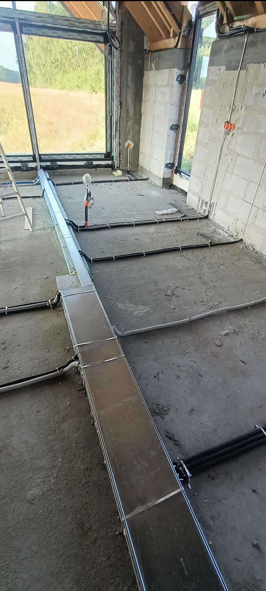 Wnętrze w trakcie prac: widoczne kable elektryczne i metalowe korytka kablowe ułożone na surowym betonie, duże okna z widokiem na zewnątrz, instalacja w trakcie realizacji.