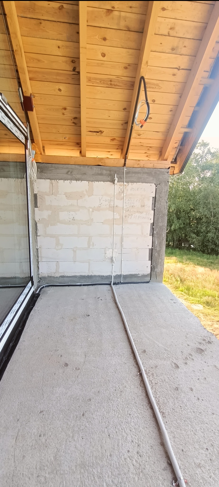 Surowy stan budynku z widoczną instalacją elektryczną na ścianie z bloczków betonowych i pod drewnianym stropem. Widoczne kable i rury instalacyjne.