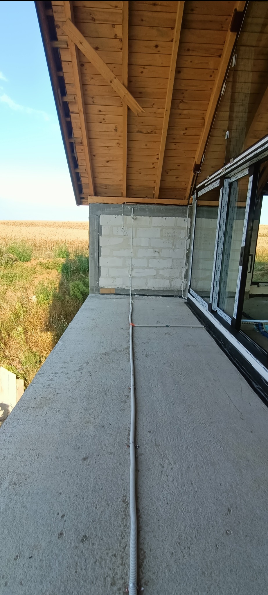 Instalacja elektryczna w peszlu na surowym balkonie z widokiem na pole. Widoczne okno przesuwne i drewniana konstrukcja dachu.