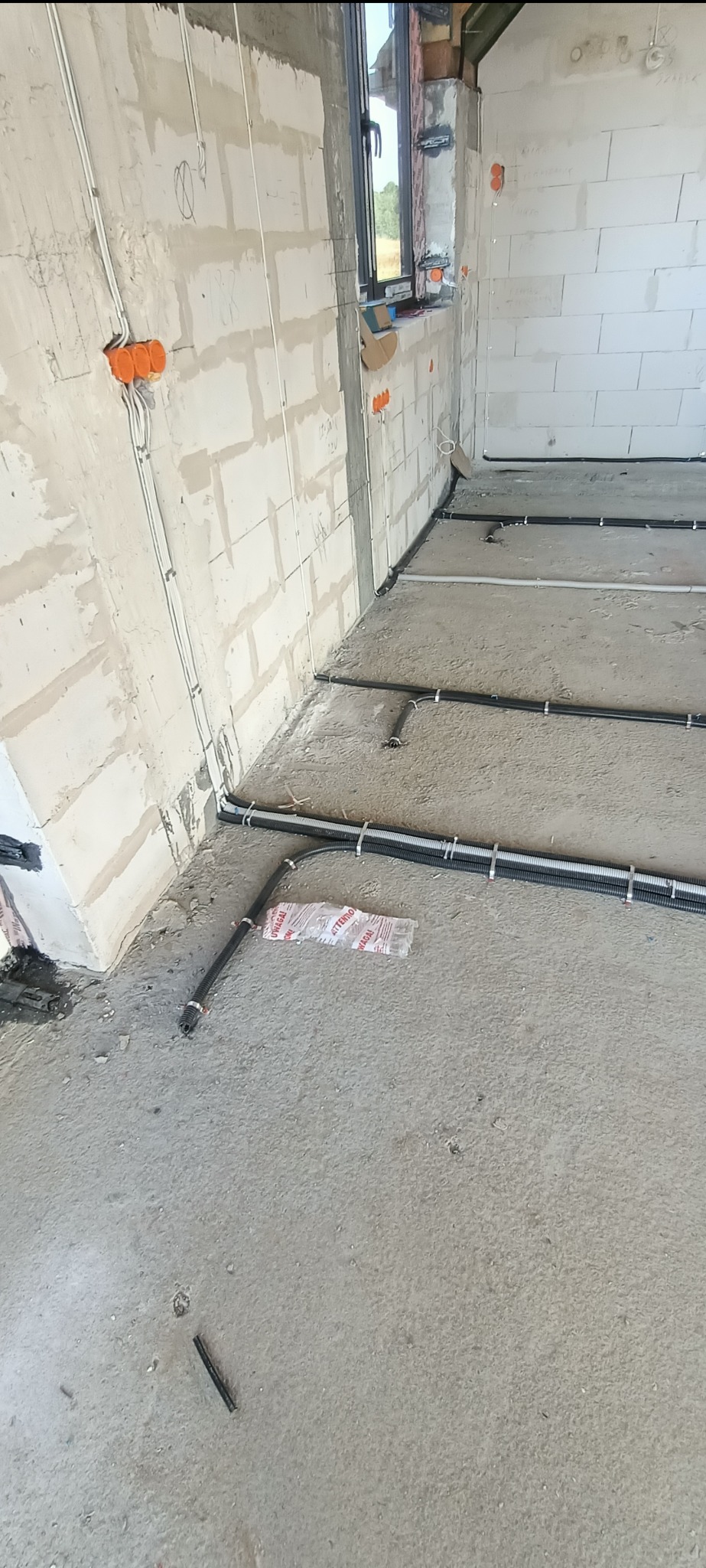 Wnętrze w trakcie prac elektrycznych: kable w peszlach ułożone na podłodze, puszki instalacyjne na ścianach z betonu komórkowego. Widoczne okno i surowy stan wykończenia.