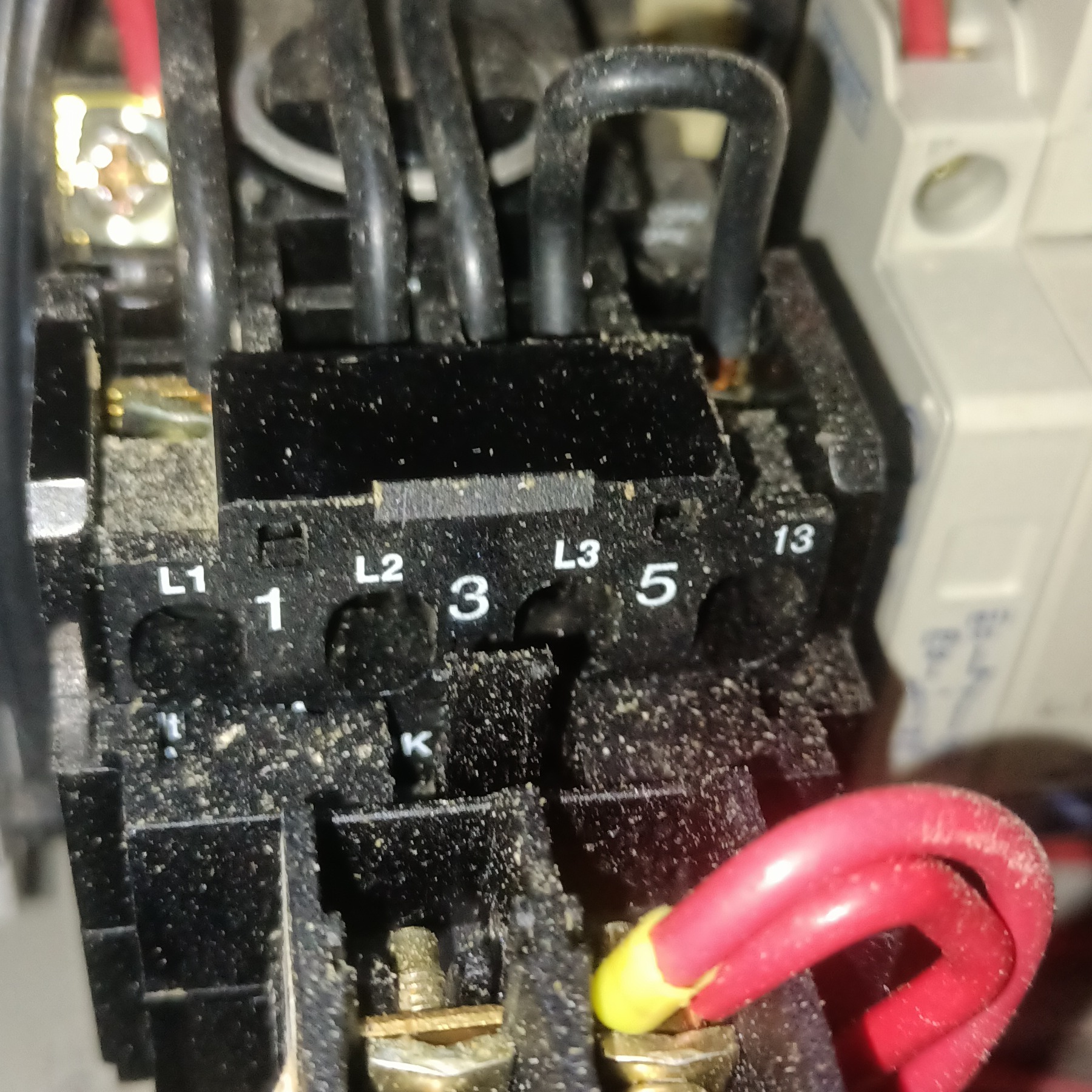 Zbliżenie na czarny stycznik elektryczny z oznaczeniami L1, L2, L3, 1, 3, 5, 13 i podłączonymi przewodami. Widoczne ślady kurzu na urządzeniu.