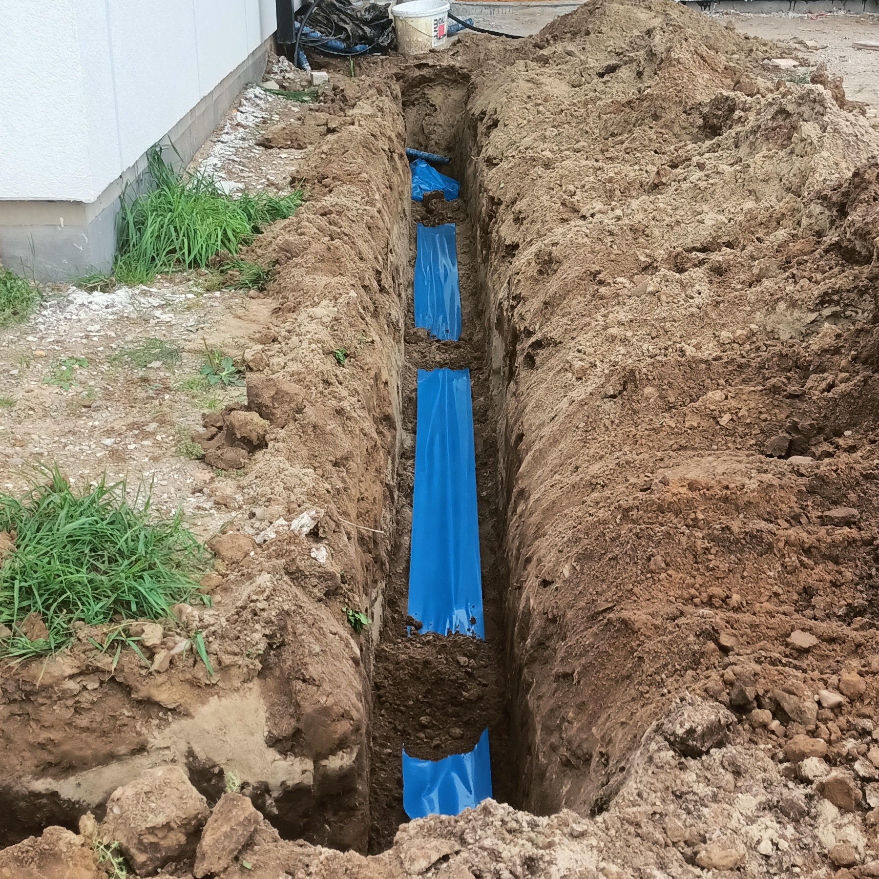 Wykop w ziemi z niebieską folią zabezpieczającą, prawdopodobnie pod instalację elektryczną. Widoczne kable i wiadro w tle. Ujęcie z góry.