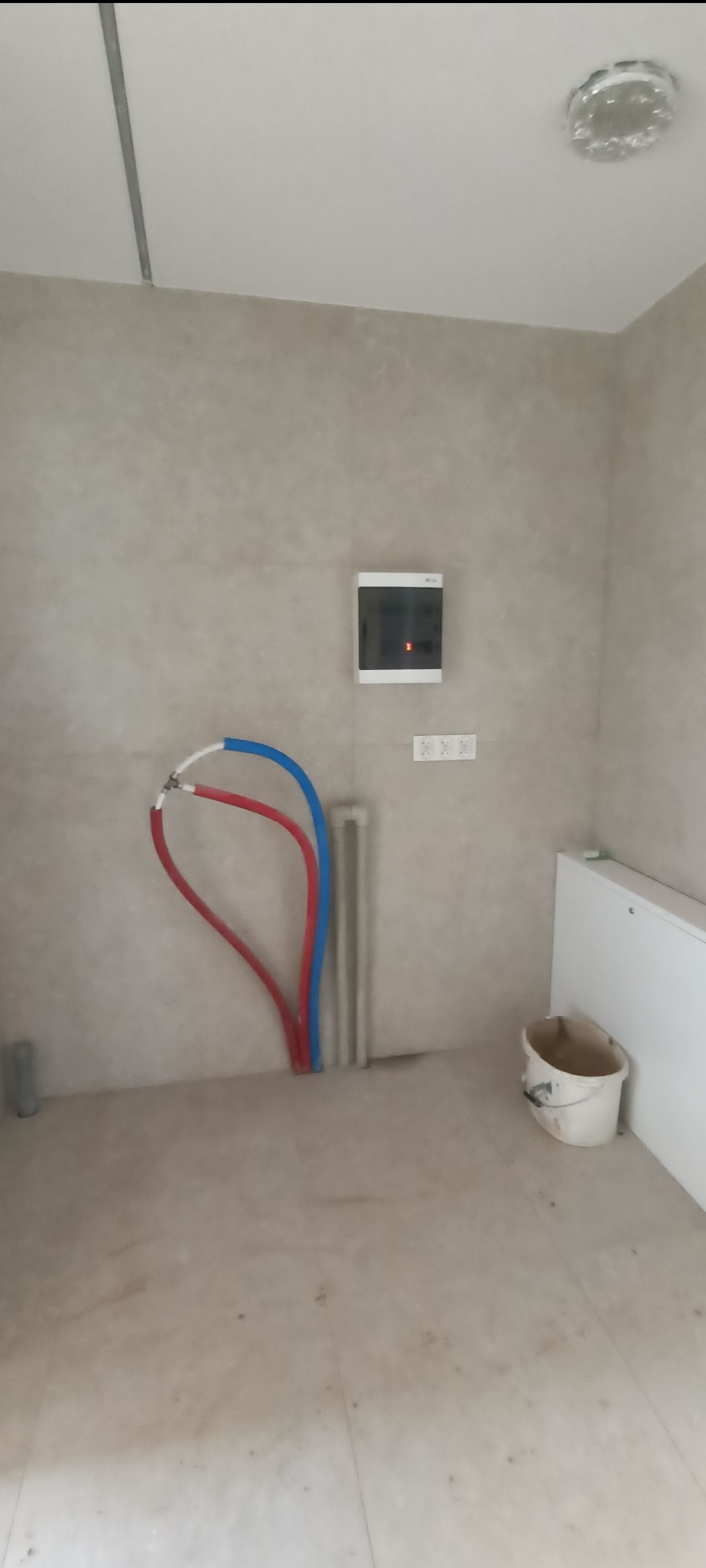 Instalacja rur wodnych (czerwona i niebieska) w pomieszczeniu z beżowymi ścianami. Widoczna skrzynka elektryczna i biały grzejnik. Podłoga wyłożona płytkami.