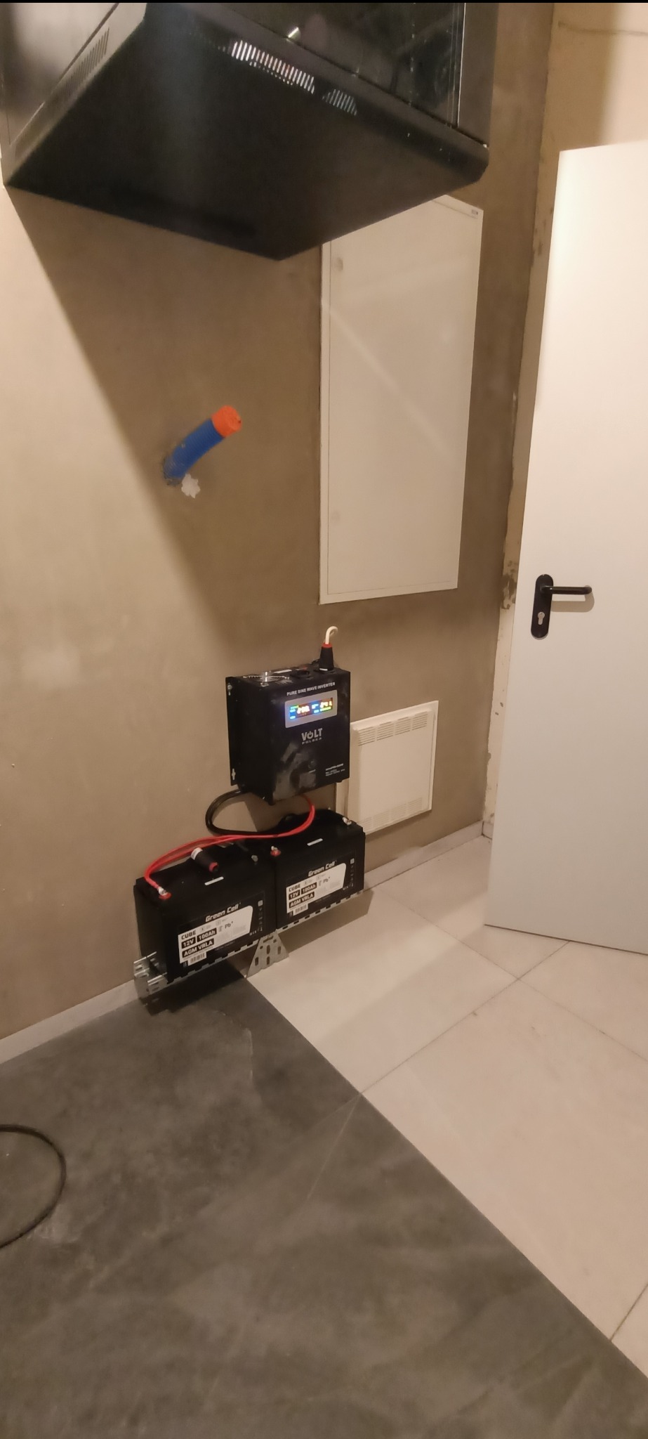 Zestaw akumulatorów Green Cell Cube 12V 100Ah i inwerter Volt Polska, zamontowane na ścianie w pomieszczeniu z płytkami, widoczne kable i elementy instalacji.