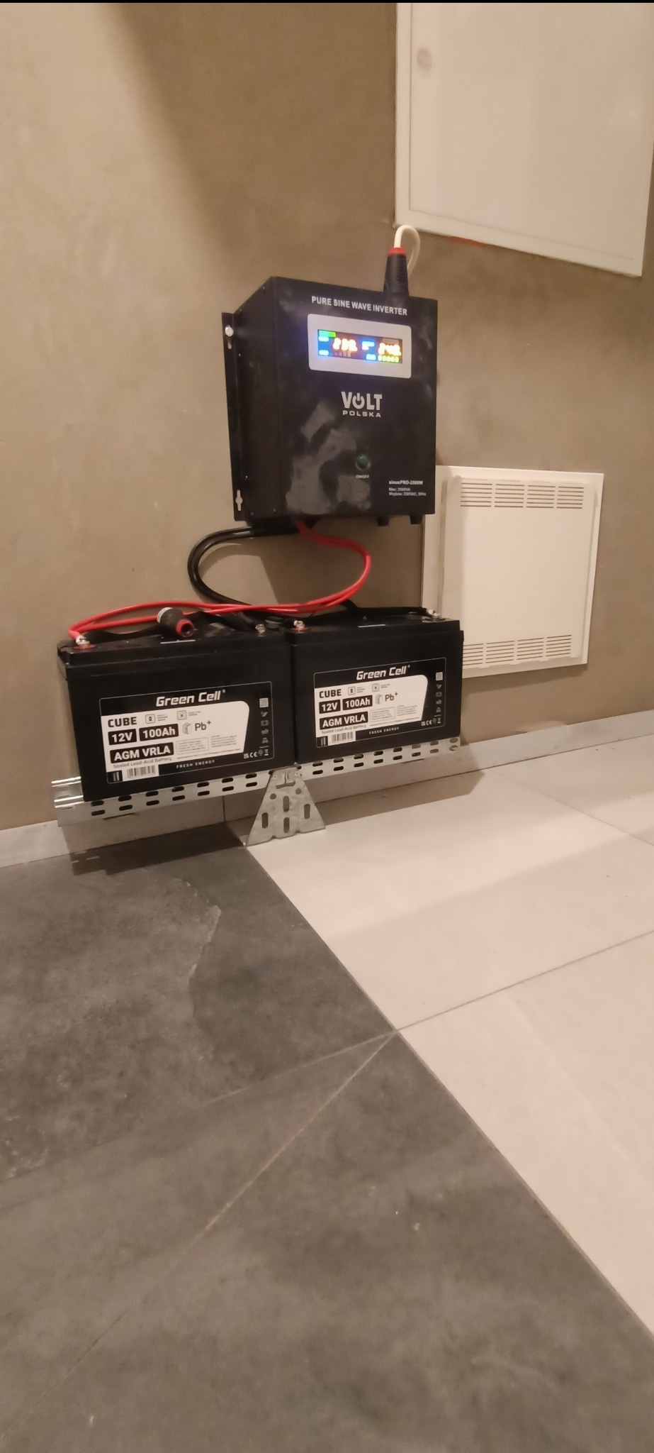 Zestaw awaryjnego zasilania: inwerter Volt Polska i dwa akumulatory Green Cell Cube 12V 100Ah, zamontowane na szynie, przy ścianie z kratką wentylacyjną.