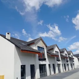 Rząd nowo wybudowanych, białych domów z ciemnymi dachami i oknami pod błękitnym niebem z chmurami. Część elewacji w stanie surowym, widoczne niedokończone ocieplenie.