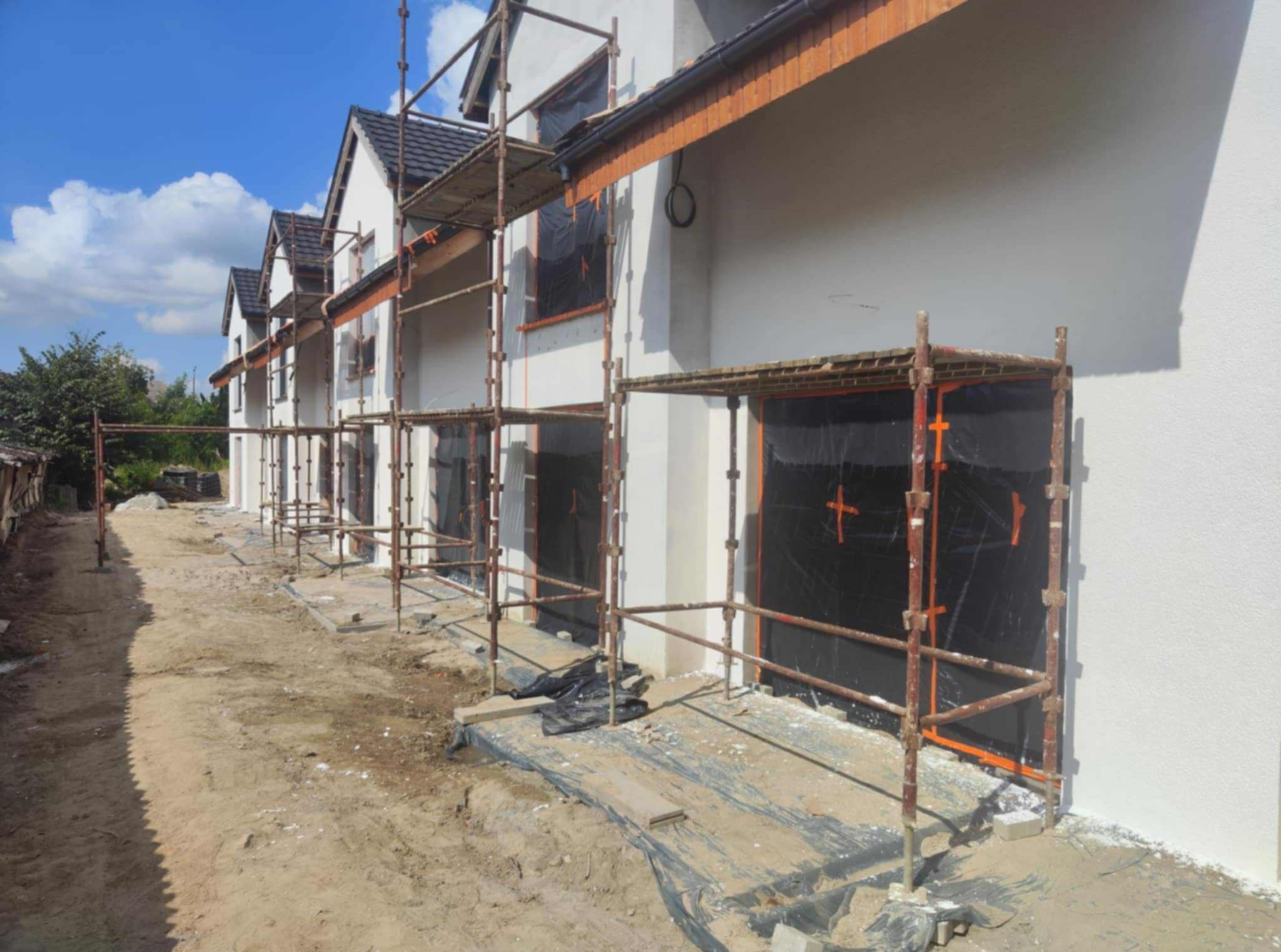 Ocieplony budynek w trakcie prac wykończeniowych. Widoczne rusztowania i zabezpieczone okna folią. Świeżo otynkowana elewacja w jasnym kolorze.