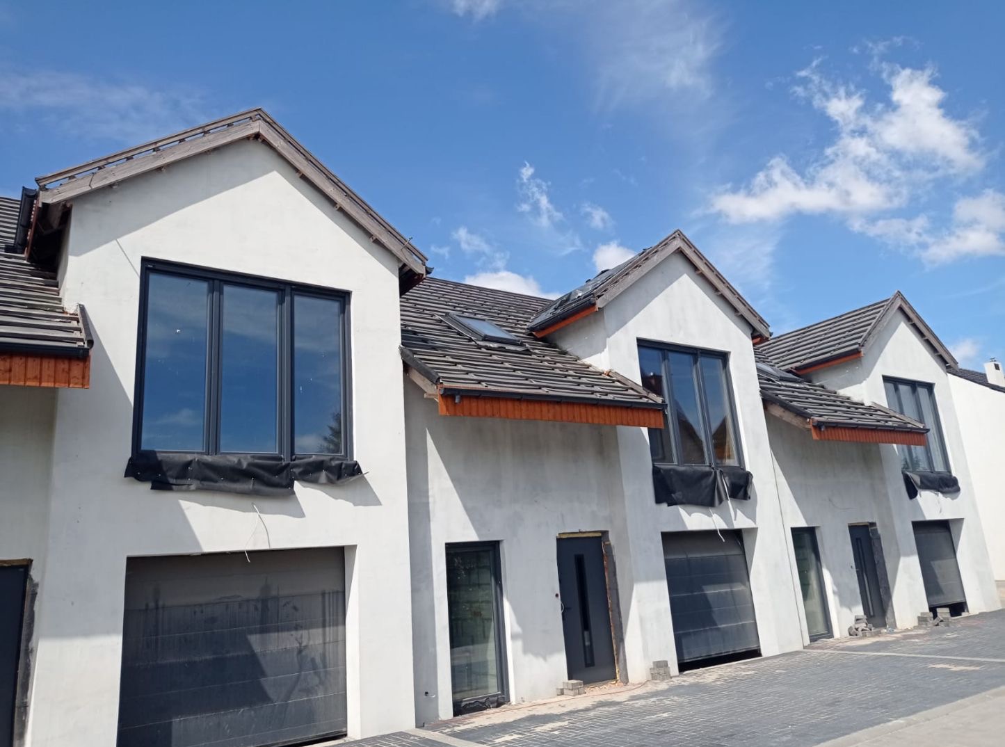Surowe elewacje szeregowców z czarnymi oknami i bramami garażowymi w słoneczny dzień. Widoczne przygotowania do dalszych prac wykończeniowych.