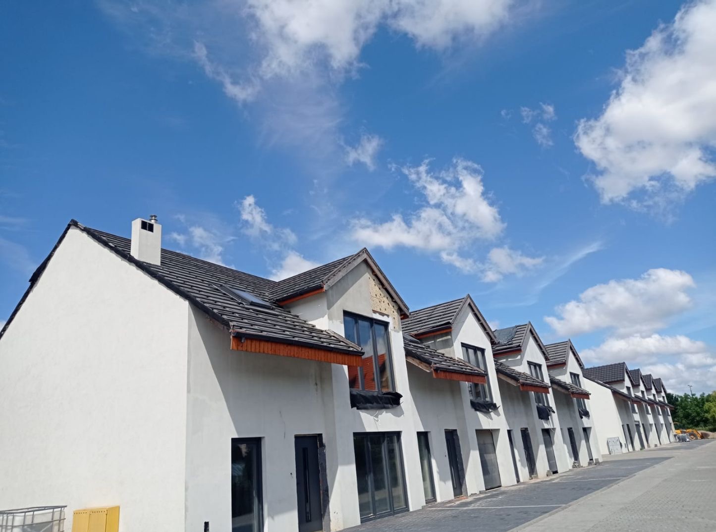 Rząd nowo wybudowanych, białych domów z ciemnymi dachami i oknami pod błękitnym niebem z chmurami. Część elewacji w stanie surowym, widoczne niedokończone ocieplenie.