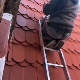 Dekarstwo Blacharstwo Sebastian Karliński JDG - Pracownik na drabinie, montujący dachówkę karpiówkę w kolorze ceglastym, widok z dołu na fragment dachu i osobę w roboczym ubraniu.