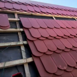 Montaż bordowej dachówki karpiówki na drewnianej konstrukcji dachu, widoczne fragmenty membrany dachowej i niezamontowane płytki.