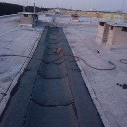 Dekarstwo Blacharstwo Sebastian Karliński JDG - Widok z góry na płaski dach z betonową rynną odwadniającą, w której leży zwinięty wąż oraz palnik dekarski, widoczne kominki wentylacyjne i odległe budynki w tle, wykonane w słabym oświetleniu.