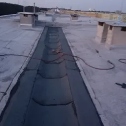 Widok z góry na płaski dach z betonową rynną odwadniającą, w której leży zwinięty wąż oraz palnik dekarski, widoczne kominki wentylacyjne i odległe budynki w tle, wykonane w słabym oświetleniu.