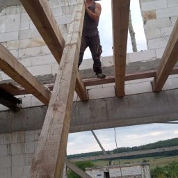 Dekarstwo Blacharstwo Sebastian Karliński JDG - Konstrukcja drewniana więźby dachowej w trakcie montażu na murowanym budynku, widok z dołu na pracownika w roboczych ubraniach, krajobraz w tle.