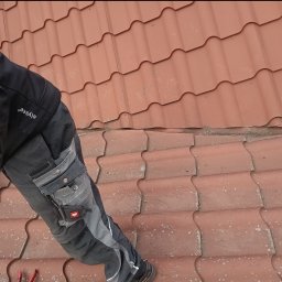 Dekarstwo Blacharstwo Sebastian Karliński JDG - Fragment dachu z czerwonej dachówki ceramicznej, na którym stoi dekarz w roboczych spodniach z widocznymi narzędziami w kieszeniach oraz butami roboczymi. Dachówka jest lekko zabrudzona. Widoczny...